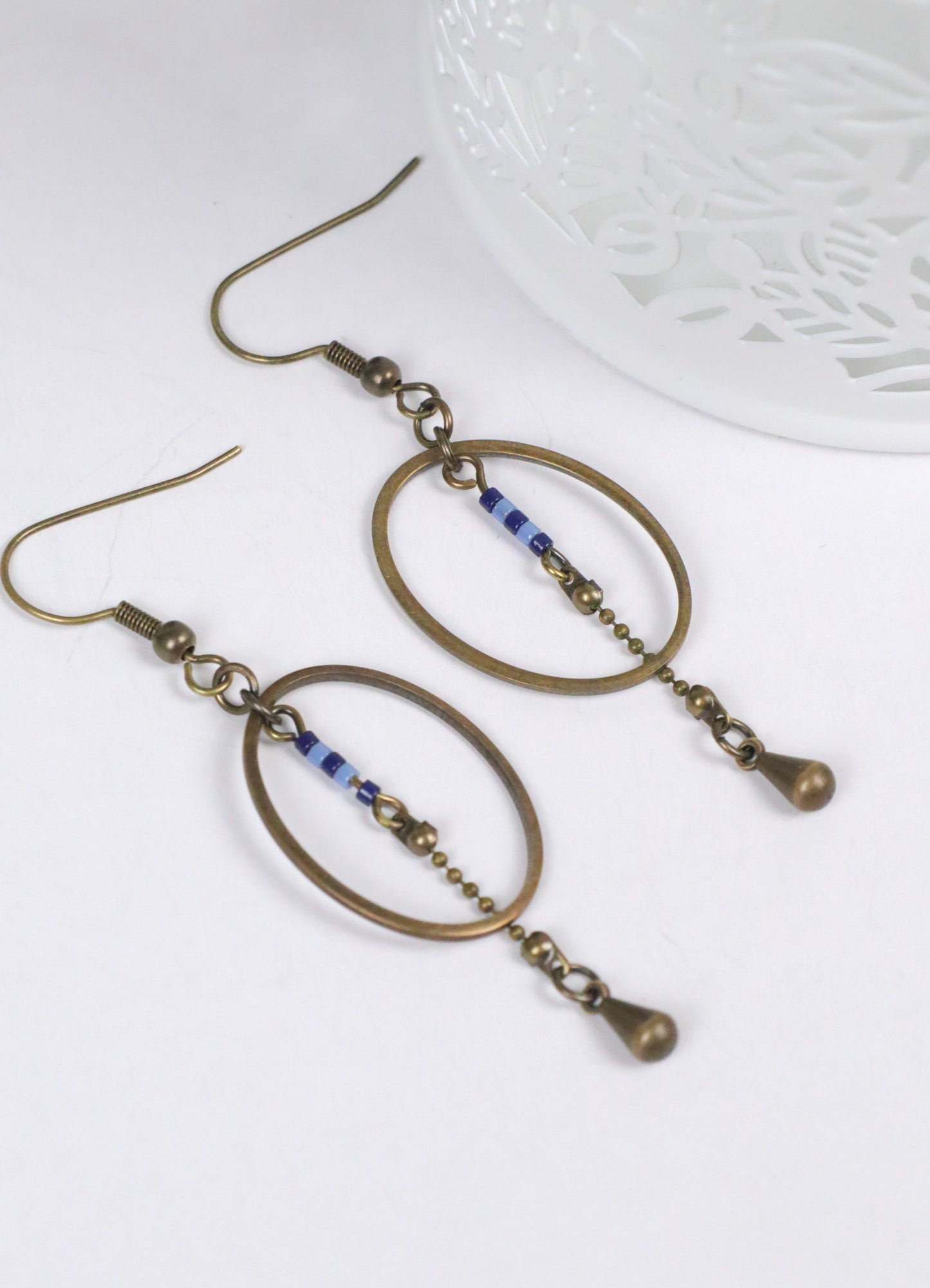 Gros plan des boucles d'oreilles pendantes, modèle Alana, ovales en laiton avec des perles bleues, à plat sur un fond blanc