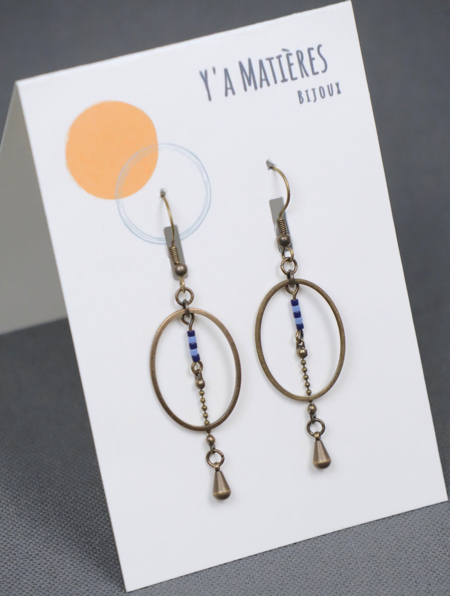Gros plan des boucles d'oreilles pendantes, modèle Alana, ovales en laiton avec des perles bleues accrochées sur une carte avec le logo Y'a Matières